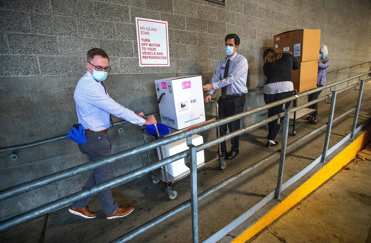 University of Washington Medical Center Montlake campus pharmacy administration resident Derek Pohlmeyer (left) and UWMC pharmacy director Michael Alwan transport a box containing Pfizer-BioNTech COVID-19 vaccines toward a waiting vehicle headed to the UW Medical Center’s other hospital campuses on Monday in Seattle. (Mike Siegel/The Seattle Times via media pool)